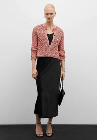 Gebreide cardigan in roze en wit met een cropped ontwerp, gedragen over een zwarte aansluitende jurk. Het model houdt een zwarte handtas vast.