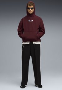 Sudadera con capucha en marrón con el logo de BMW Motorsport, bolsillo de tipo canguro en el frente. Pantalones anchos negros con cadenas, combinados con zapatos negros.