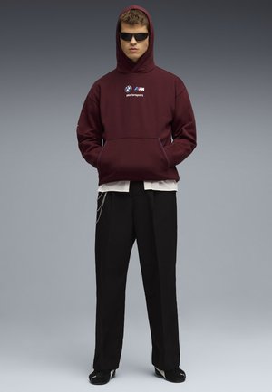 Bordeaux hættetrøje med BMW Motorsport-logo, frontlomme. Sorte bredbenede bukser med kæder, parret med sorte sko.