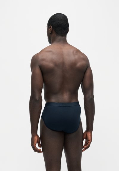 Homme debout de dos, portant un slip bleu foncé devant un fond clair uni.