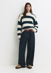 Donna che indossa un maglione a righe bianche e blu navy, jeans larghi scuri con una cintura rossa e stivali da trekking beige, in piedi contro un muro bianco.
