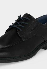 Chaussure formelle en cuir noir avec laçage fermé, détails cousus et talon bas sur fond clair uni.