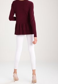 Bordowy sweter z długimi rękawami i plisowanym dołem, w połączeniu z białymi obcisłymi spodniami i beżowymi butami na wysokim obcasie, widziany od tyłu.