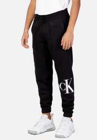 Pantalones de chándal negros con cintura ajustable, bolsillos laterales y un logo blanco de Calvin Klein en la parte inferior de la pierna. Combinados con zapatillas blancas.