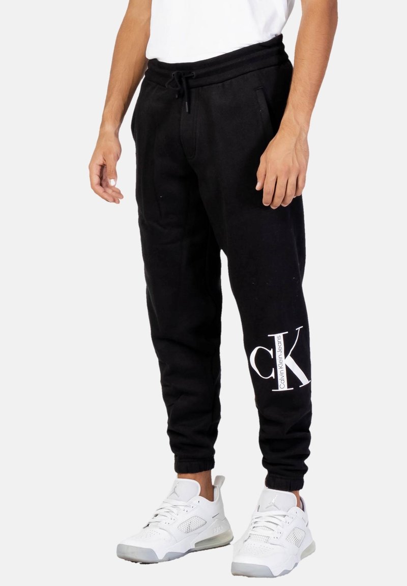 Pantalones de chándal negros con cintura ajustable, bolsillos laterales y un logo blanco de Calvin Klein en la parte inferior de la pierna. Combinados con zapatillas blancas.