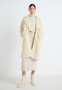Krämfärgad fuskpälsjacka med slagkrage, över en beige ribbad crop top, beige kjol och cremfärgade ankelskors med dragkedja.
