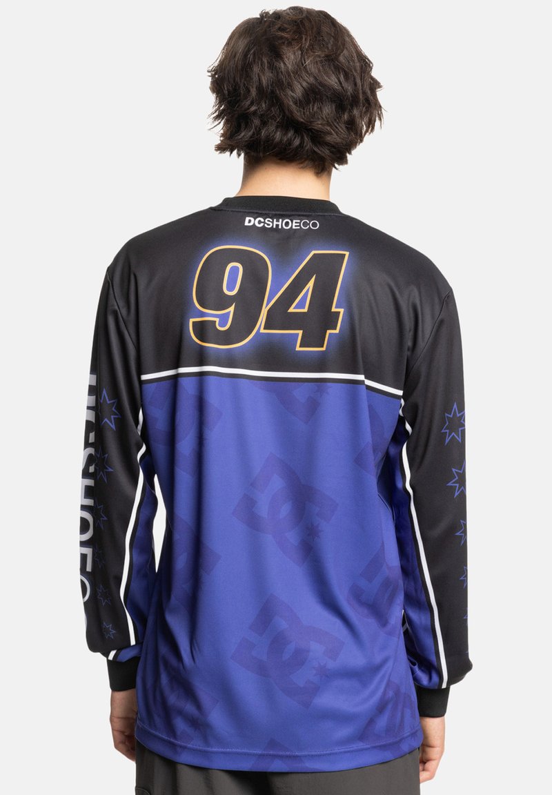 Dolgi rokavi dres v črni in purpurni barvi z belimi črtami, z velikim rumenim številom "94" na hrbtu in vzorci DC logotipa.