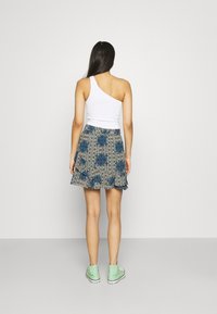 Haut blanc texturé à une épaule associé à une jupe courte à motifs floraux bleu et beige, agrémentée d'un ourlet à volants et de baskets vertes.