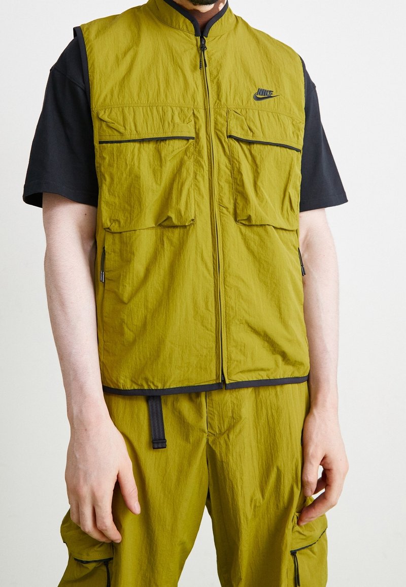 Man som bär en olivgrön ärmlös zip-up väst och matchande cargo-byxor över en svart t-shirt med kort ärm, som står mot en enfärgad bakgrund.