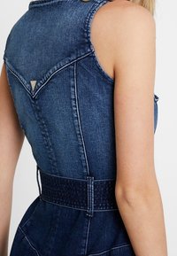 Denimmekko, jossa on istuva yläosa, hihaton muotoilu ja vyötäröosa. Tumansininen kangas, jossa on kontrastiväriset ompeleet ja takana kolmio-detaili.
