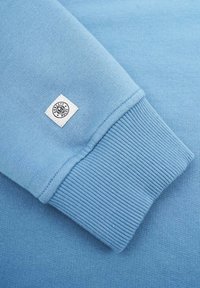 Vaaleansininen huppari, jossa on ribattu hihansuu. Materiaali näyttää sileältä ja hihansuussa on pieni valkoinen tagi, jossa on "Honesty Rules" -logo.