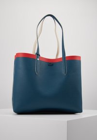 Niebieska torba tote z teksturowanej skóry z czerwoną górną lamówką, z dwoma kremowymi uchwytami i eleganckim, minimalistycznym wzornictwem.