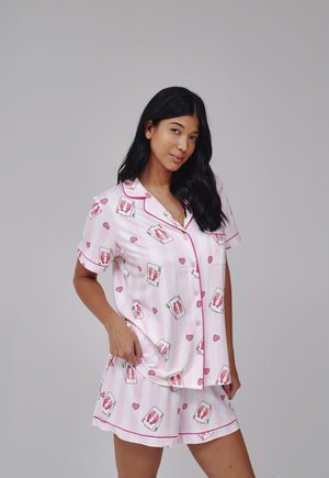 Vrouw met lang zwart haar, gekleed in een roze pyjamashirt met hartjes- en kaartpatroon en bijpassende korte broek, tegen een grijze achtergrond.