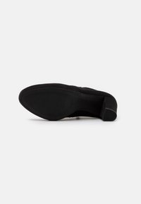 Zapato bajo de ante negro con tacón bajo, acostado de lado, mostrando la suela texturizada y el tacón cuadrado sobre un fondo blanco liso.