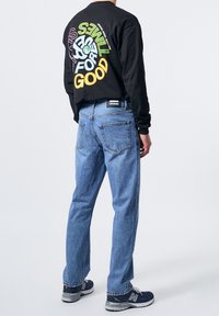 Homme portant une chemise noire à manches longues avec un texte circulaire coloré et un jean bleu clair, vu de dos, debout sur un fond blanc uni.