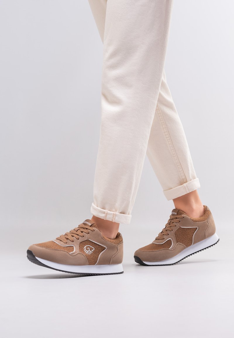 Persoon die beige broek en bruine suède sneakers met witte zolen draagt, staand op een lichtgrijze ondergrond tegen een effen achtergrond.