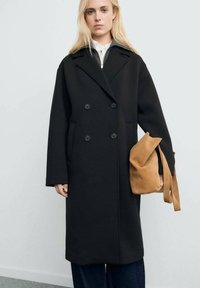 Abrigo negro de doble botonadura con grandes solapas y dos bolsillos frontales. Lleva una parte superior blanca y lo combina con un bolso de ante color marrón. Ajuste casual.