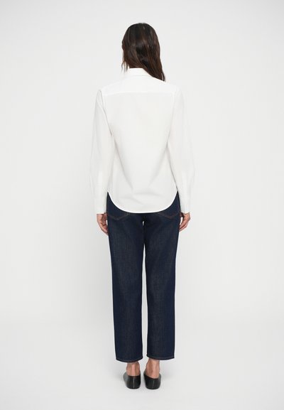 Camicia bianca a maniche lunghe con bottoni e orlo curvo, abbinata a jeans dritti blu scuro. Scarpe nere basse completano il look.