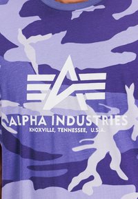 Lila kamuflažna majica ima okrugli izrez, kratke rukave in bel logo "Alpha Industries" z geometrijskimi akcenti.