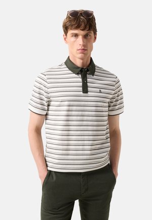 Ung mand med solbriller på hovedet, en hvid og sort stribet polo med mørkravet og mørkegrønne bukser med hænderne i lommerne.