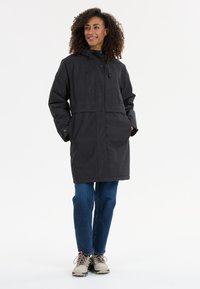 Parka nera impermeabile con cappuccio regolabile, bottoni a pressione e tasche frontali profonde, indossata sopra jeans blu e scarpe da trekking beige.