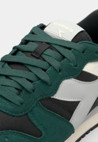 Grønn og svart semsket skinn- og mesh-sneaker med kontrastfulle grå detaljer, teksturerte sko-lisser og bølgete design på overdelen nær en hvit logo.