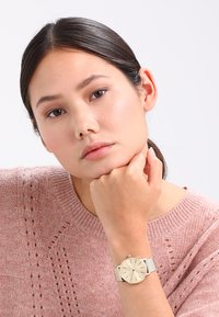 Gold-tone watch with minimalist round face, silver mesh band, and quartz movement; worn on a wrist resting against a chin.