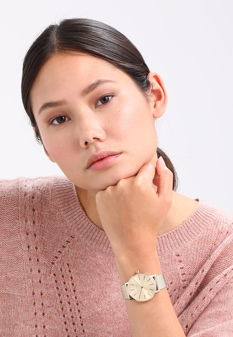 Gold-tone watch with minimalist round face, silver mesh band, and quartz movement; worn on a wrist resting against a chin.
