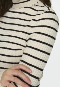 Pull à col roulé côtelé en crème avec des rayures horizontales noires. Dispose de poignets ajustés et d'une coupe près du corps pour la chaleur et le confort.