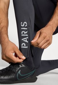 Sorte treningsleggings med "PARIS" i reflekterende grå. Leggingsene har en sidelynlåsdetalj. En svart Nike-sko er synlig.