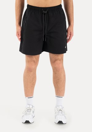 Mand iførende sorte shorts med snøre, hvide mærkesokker og hvide sportssko, står med hænderne afslappet ved siderne.