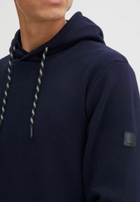 Homme portant un sweat à capuche bleu marine avec des cordons de tirage noir et blanc et une petite pièce noire sur le haut de la manche.