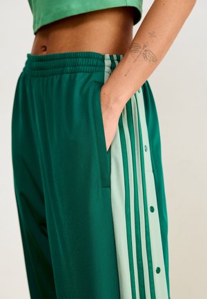 Person trägt grüne Trainingshose mit hellgrünen Seitenstreifen und Druckknöpfen, Hand in der Tasche, zeigt ein Schmetterlingstattoo am Unterarm.