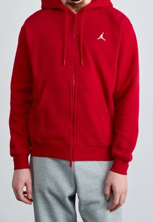 Mand iført en rød hættetrøje med lynlås med en hvid Jumpman-logo på brystet og grå joggingbukser mod en ensfarvet baggrund.