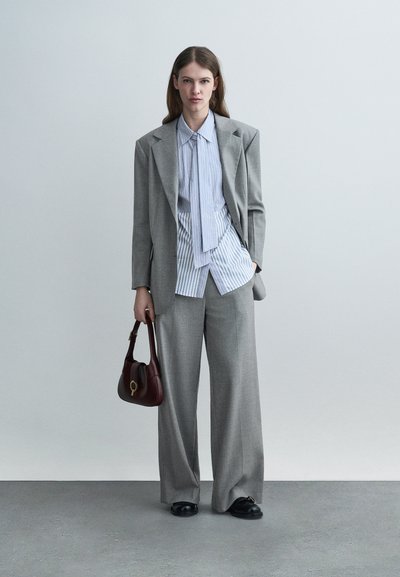 Femme portant un costume gris surdimensionné avec une chemise rayée ample, tenant un sac à main marron foncé, debout sur un sol gris devant un mur clair uni.