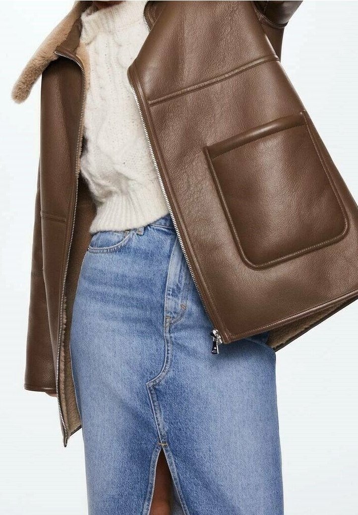 Giacca di pelle marrone con colletto in pelliccia, caratterizzata da due grandi tasche frontali, abbinata a una gonna in denim azzurro chiaro con uno spacco frontale.