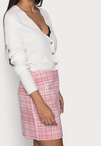 Bílý žebrovaný cardigan s hlubokým výstřihem do V a zlatými knoflíky, v kombinaci s růžovou kostkovanou mini sukni s texturovaným vzorem.