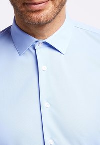 Hemelsblauw overhemd met een spread collar, zachte textuur en witte knopen langs de sluiting, met een getailleerde pasvorm.