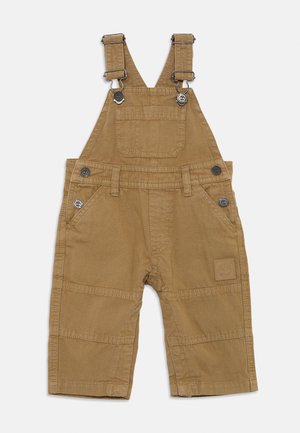 Salopette en coton beige avec des bretelles métalliques ajustables, des poches avant et latérales, et un patch logo carré sur la jambe. Texture lisse, design utilitaire.