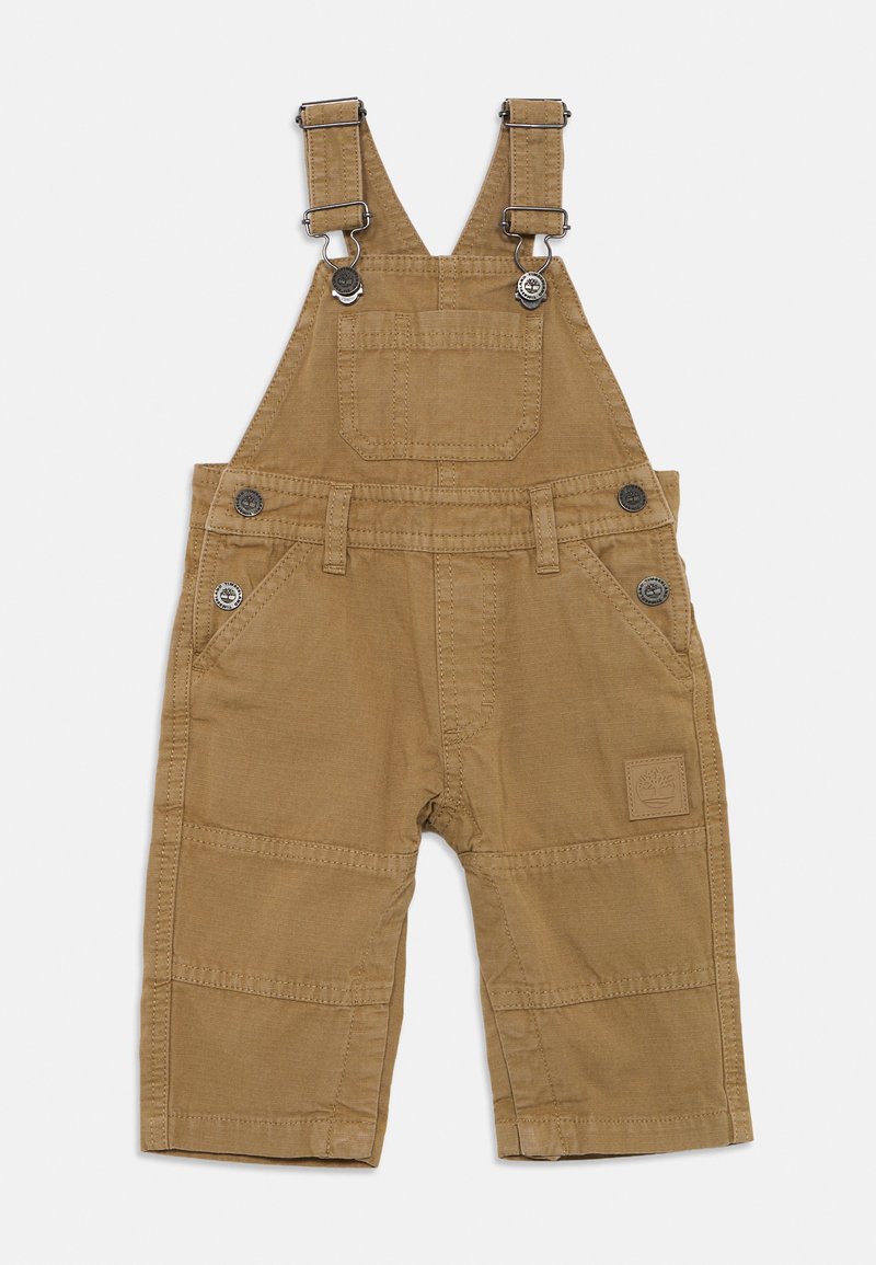 Brune bomullshelter med justerbare metallstropper, front- og sidelommer, samt en firkantet logopatch på benet. Glatt tekstur, bruksvennlig design.