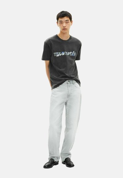 Juodas trumpomis rankovėmis marškinėliai su daugiaspalviu grafiniu tekstu priekyje, derinami su šviesiai pilkais tiesiais džinsais ir juodais batais.