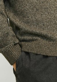Pull en tricot de couleur vert olive foncé, avec une surface texturée, des poignets et un ourlet côtelés, associé à un pantalon en tissu noir.