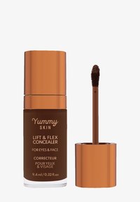 Correcteur liquide marron dans un flacon en verre cylindrique haut, avec un capuchon en cuivre brillant. Comprend un applicateur en forme de wand avec un embout duveteux.