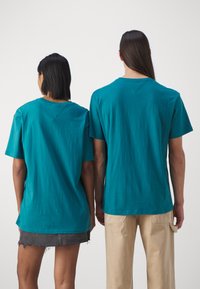 Teal puuvillased t-särgid lühikeste varrukatega, nähtavad õmblused ja mugav lõige. Kahte indiviidi on nähtud tagantvaates, üks neist denimseelikus.