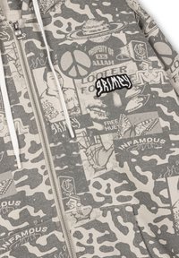 Sudadera gris con cremallera que presenta un patrón de camuflaje con varios estampados gráficos y el texto bordado "GRIMEY" en el pecho.