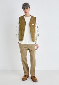 Vest îmblănit khaki peste o cămașă albă cu mânecă lungă, pantaloni bej și cizme maro cu șiret. Șapcă gri cu detaliu logo. Fundal simplu.