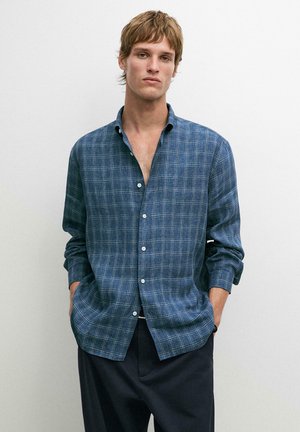Jeune homme les mains dans les poches, portant une chemise à carreaux bleue boutonnée et un pantalon foncé, sur un fond clair uni.