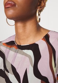 Blusa estampada multicolor com decote redondo e textura suave, acompanhada de brincos de argola dourados. As cores principais incluem roxo, castanho e verde.