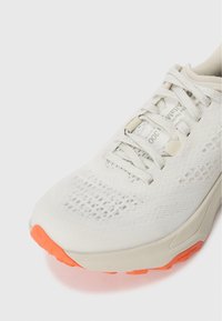 Lehká běžecká obuv s síťovinovým svrškem v krémové barvě a živě oranžovými akcenty, se strukturovaným povrchem a bezpečným uzavřením na tkaničky.