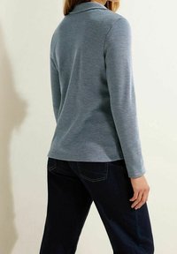 Langærmet, kravebeklædt top i lyseblå bomuldsblanding med en struktureret overflade. Båret med mørke denimbukser, vinklet set bagfra.